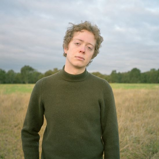 A person in a field wearing olive green sweater. The person has short hair and is staring at the camera. Behind them is a field with trees in the distance. 