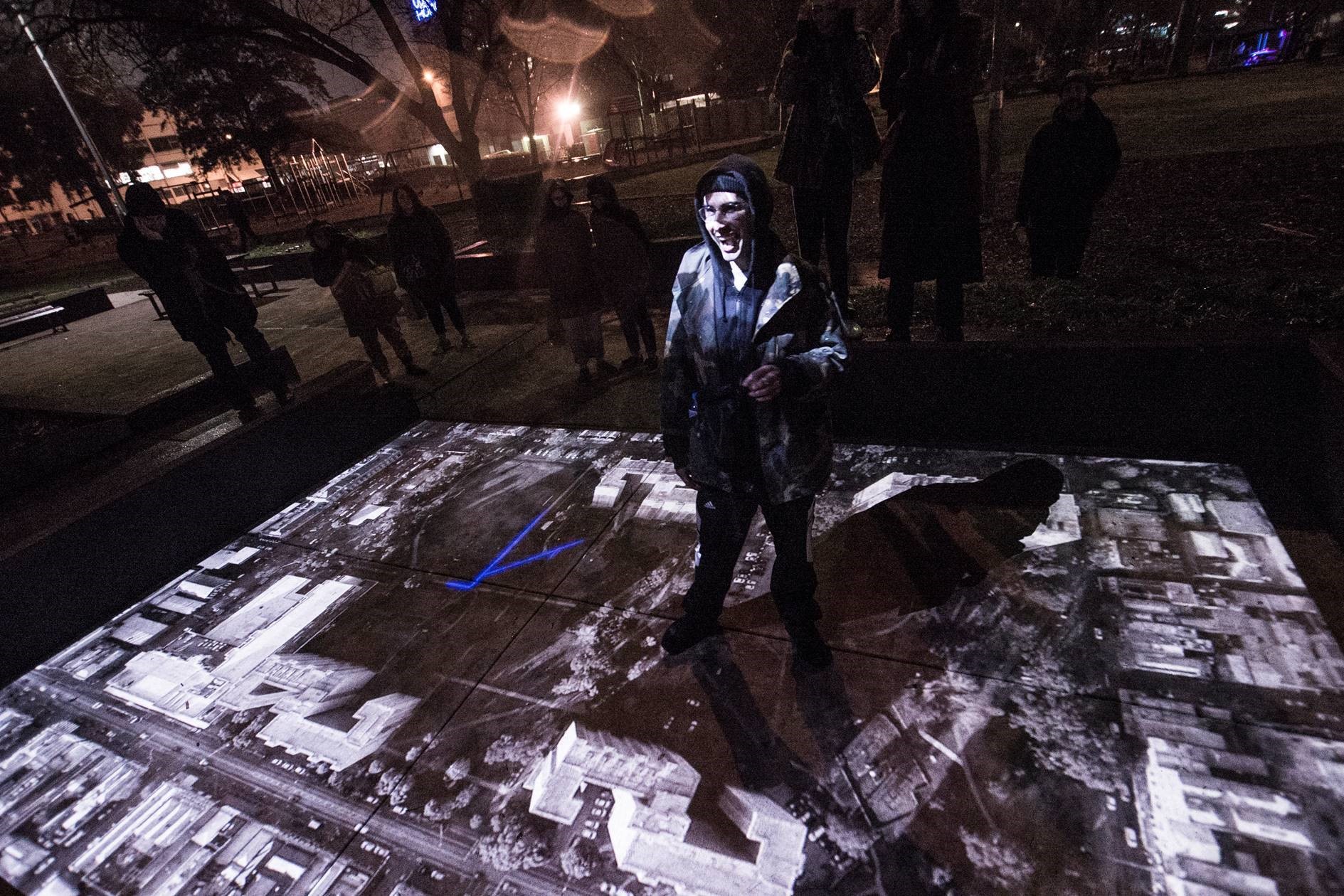Image of a person smiling, standing on top of a birds eye view of a city which is projected onto the floor. 