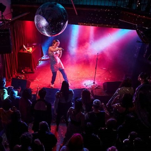 Photo of an artist coloured with pink and blue light hues, pointing to a packed out crowed, lit with the sparkle of a disco ball