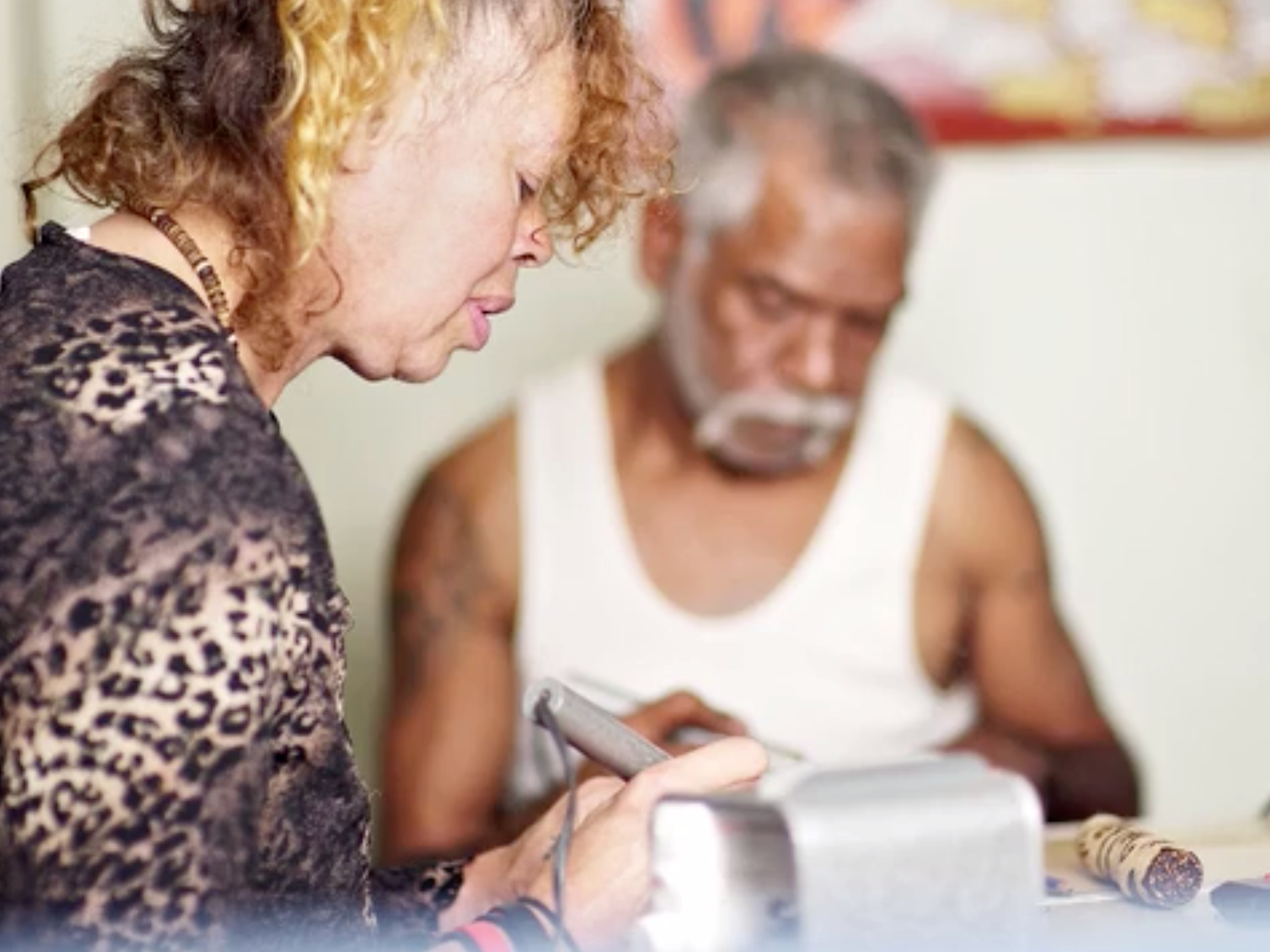 Two people, looking down towards a table where they are creating art. One person is wearing a leopard print top, and the other is wearing a white singlet. 