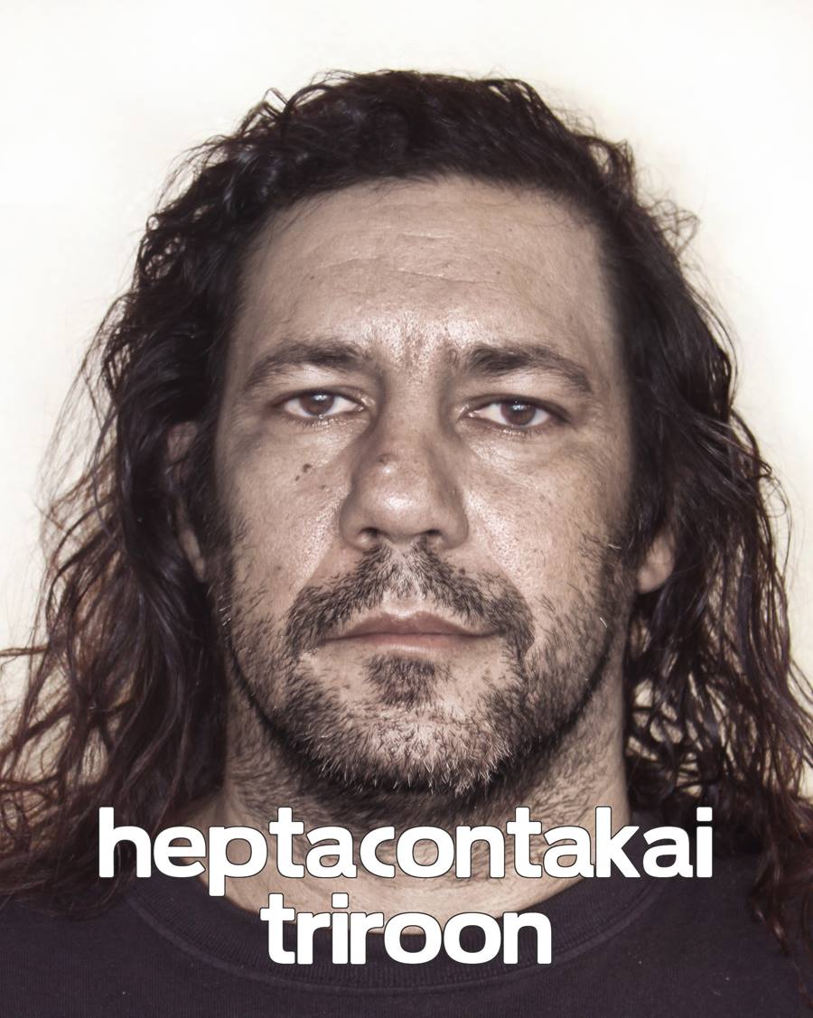 An image of a person with a beard and long brown hair with a neutral expression with 'heptacontakai triroon' written in white text over the image. 