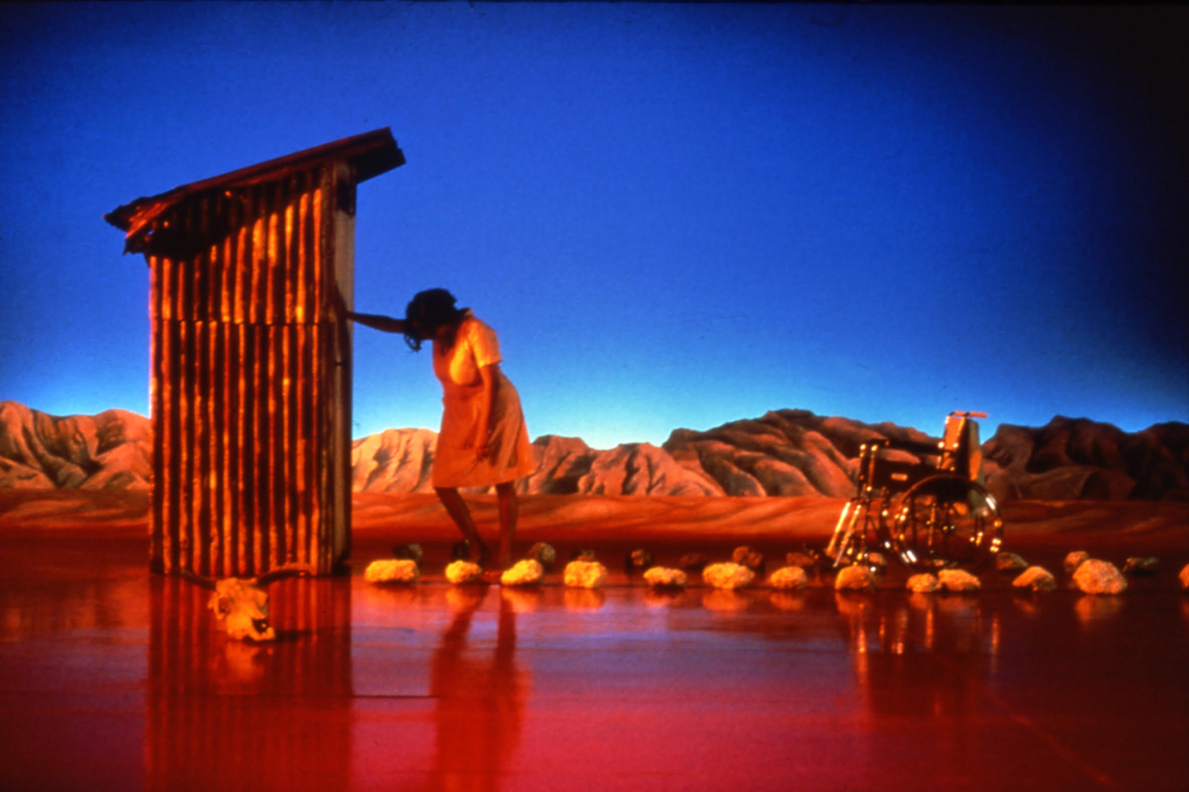 Aboriginal woman on a stage with the outback a the backdrop. She is leaning against an outhouse.