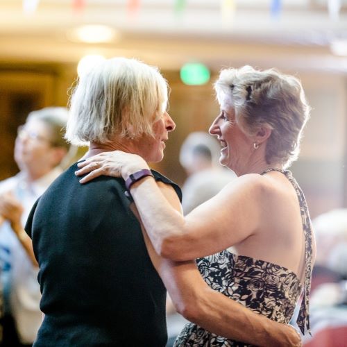 two grey haired ladies waltz together smiling inside a dance hall