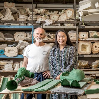 Two artists stand together in front of a table of their works: green sculpture mold, as well as a stacked cabinet of clay behind them. 