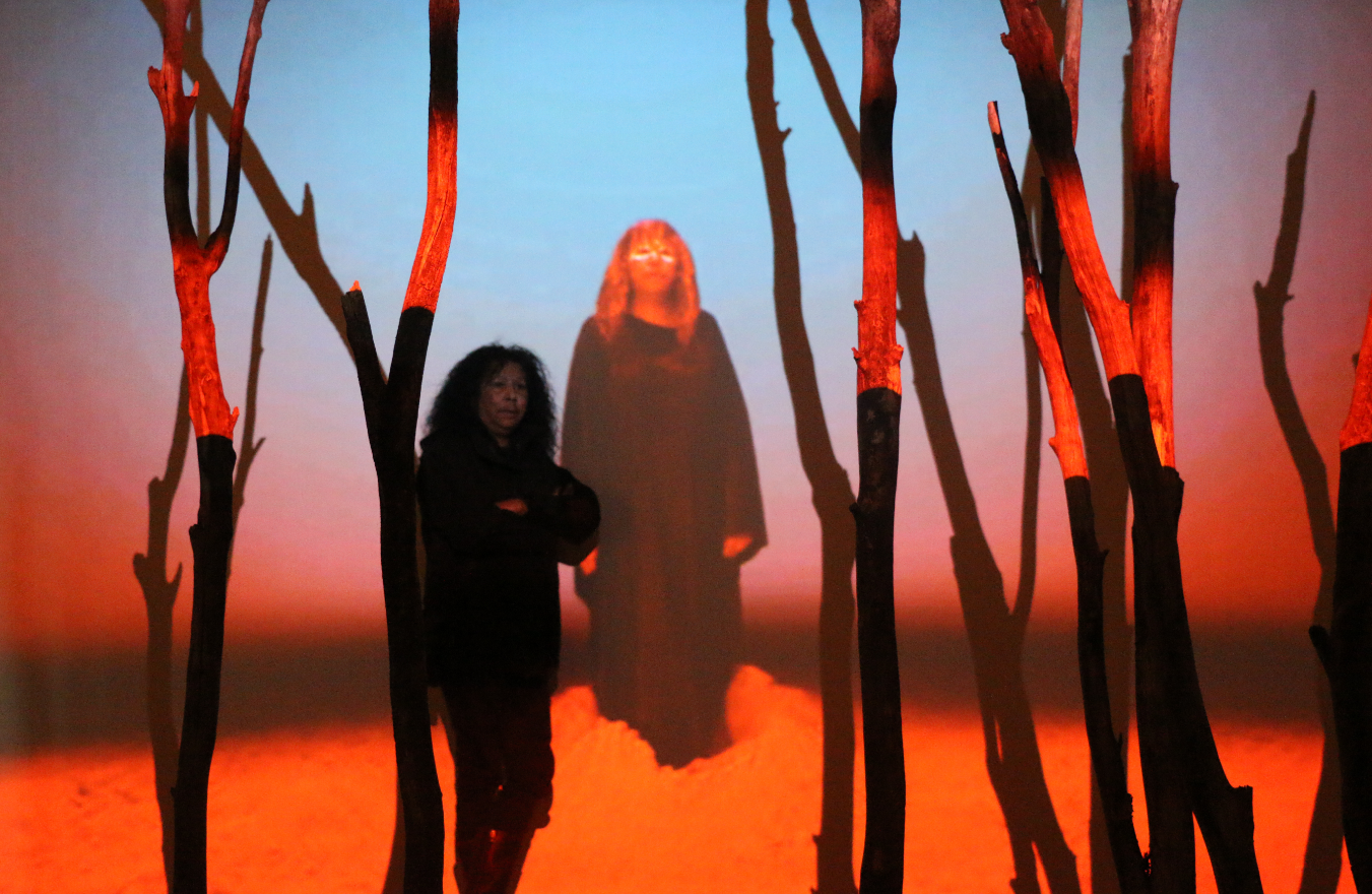 A person in a field of trees with blue and red projections over the trees. Behind the figure is a projected image of another figure standing proud.