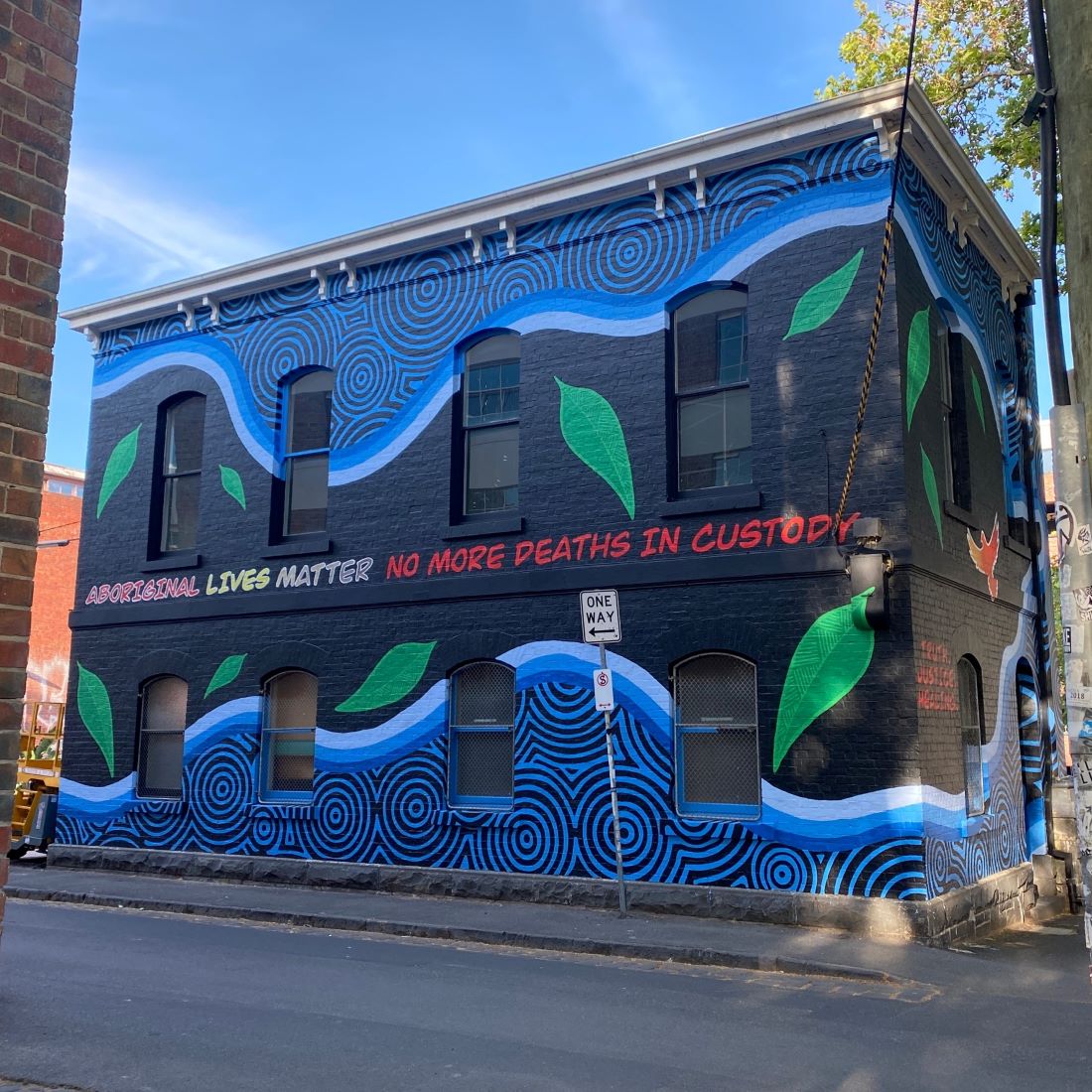 A building on a corner, which has a full-scale mural painted on it. The painting includes Manna gum leaves and the words Aboriginal Lives Matter, No more deaths in custody.