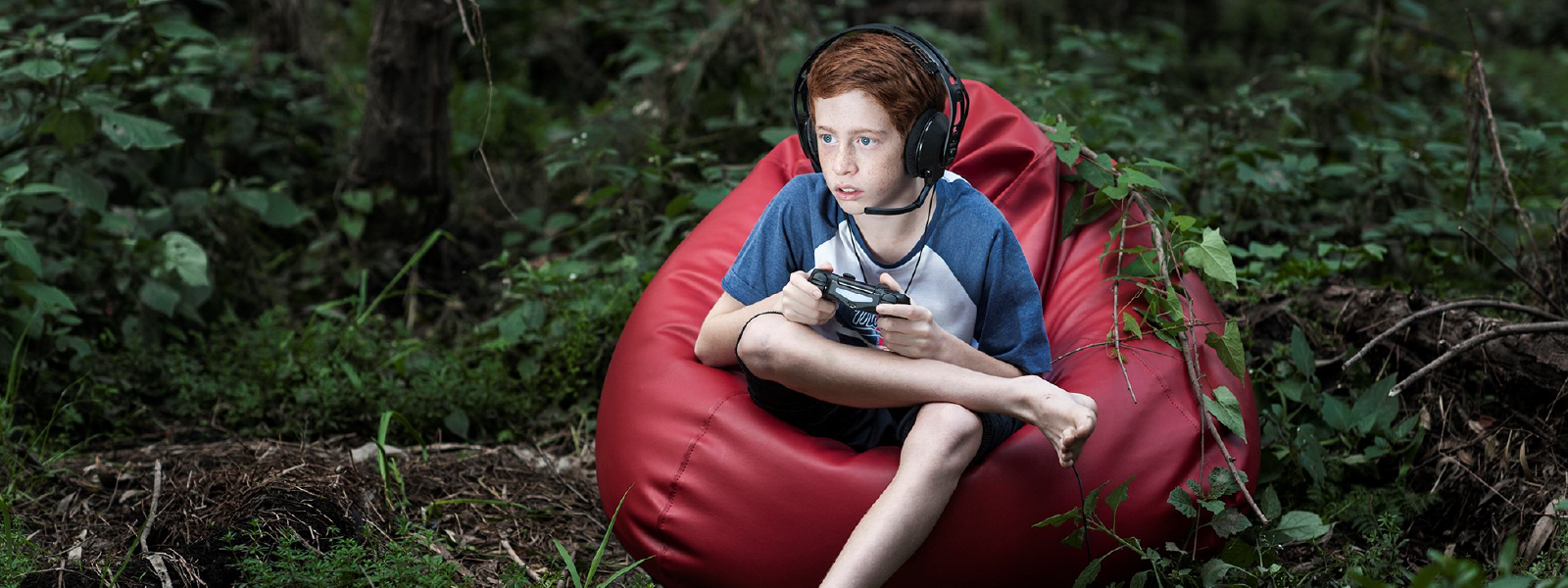 A child sits on a bright red beanbag in a forest. They are using a Playstation controller with different game cards on the forest floor around them. 