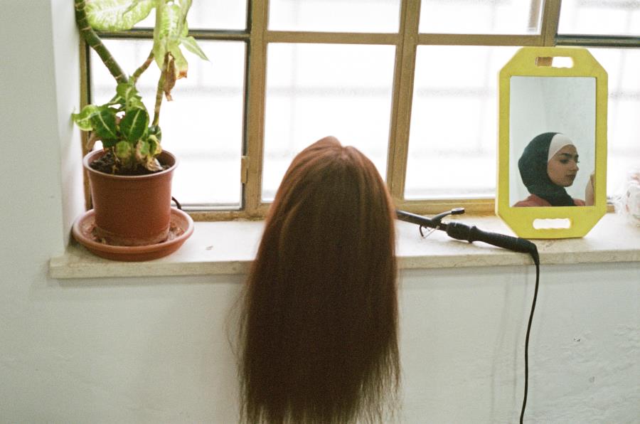 A disembodied wig on a windowsill facing a mirror with an Islamic girl reflected in it.