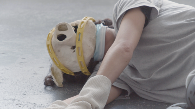Video still of the artist performing on the floor, cropped from their waist to their head. They wear a ceramic mask that resembles a pigs head and white gloves.  