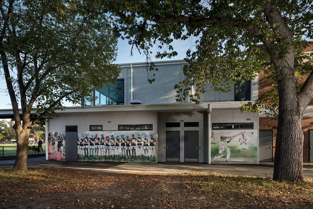 A building with a mural on it. From left to right - people running on a running track, footy players linked in arms wearing yellow and black guernseys and a cricket player in whites hitting a ball