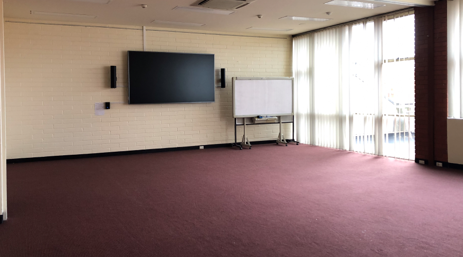 The Richmond Library Meeting Room with a wall mounted screen and whiteboard. The room is empty with no furniture set up, and sunlight is showing through the blinds.