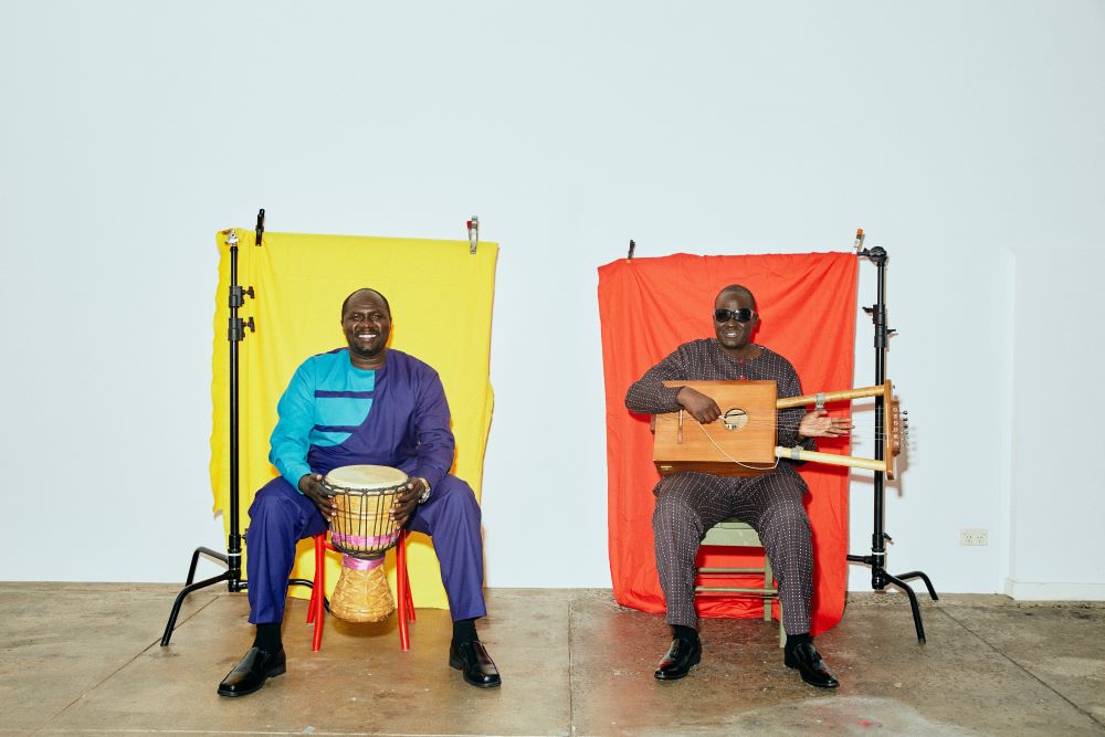 Gordon Koang and his cousin Paul Biel sitting in front of coloured sheets, playing traditional instruments. 